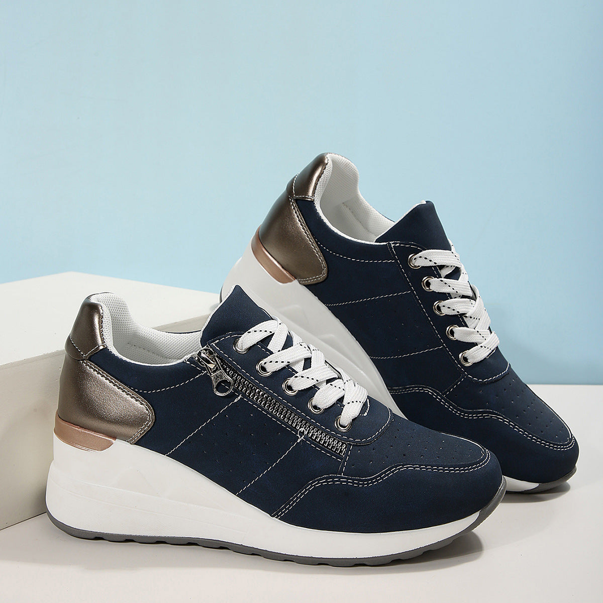 A pair of women's sneakers with lace-up closure, featuring a round toe shape, low-top design, and a flat sole. The sneakers are primarily blue with artificial PU leather and a rubber sole, set against a backdrop of a cobbled street.